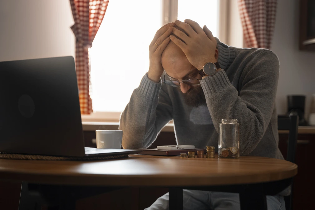 A man with a beard and glasses is sitting at a table with his head in his hands. A laptop, coffee mug, phone, coins, and a jar are on the table.