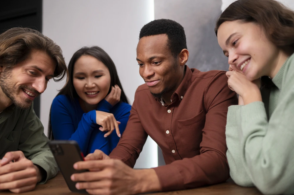 Four people are gathered around a table, looking at a smartphone held by a person in the center. They are smiling and appear to be engaged with the content on the phone. The group appears to be diverse, possibly of different ethnicities.