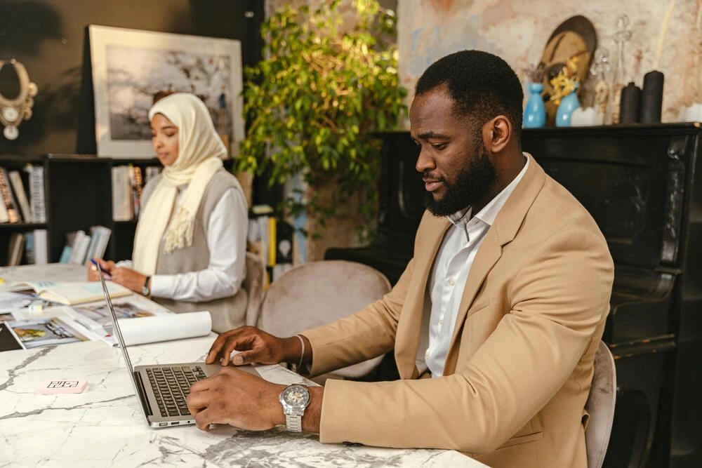 A man with dark hair and a beard is working on a laptop at a desk. In the background, a person wearing a hijab is also working at the desk. The room appears to be an office or creative space with artwork and plants.