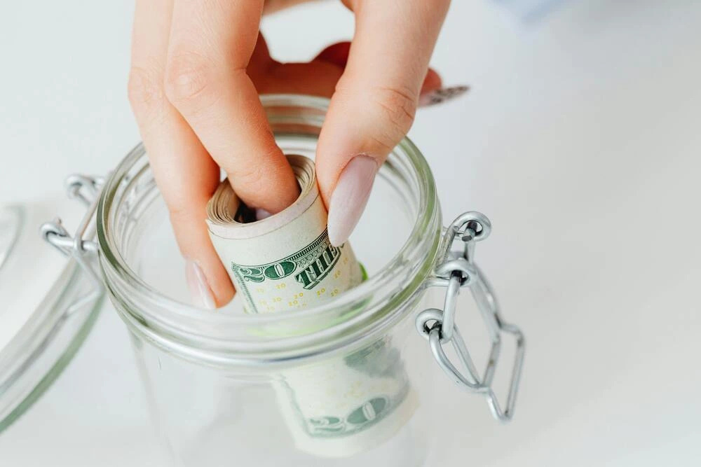 A close-up shot of a hand with pale pink nails placing a roll of US $20 bills into a clear glass jar with a metal clasp.