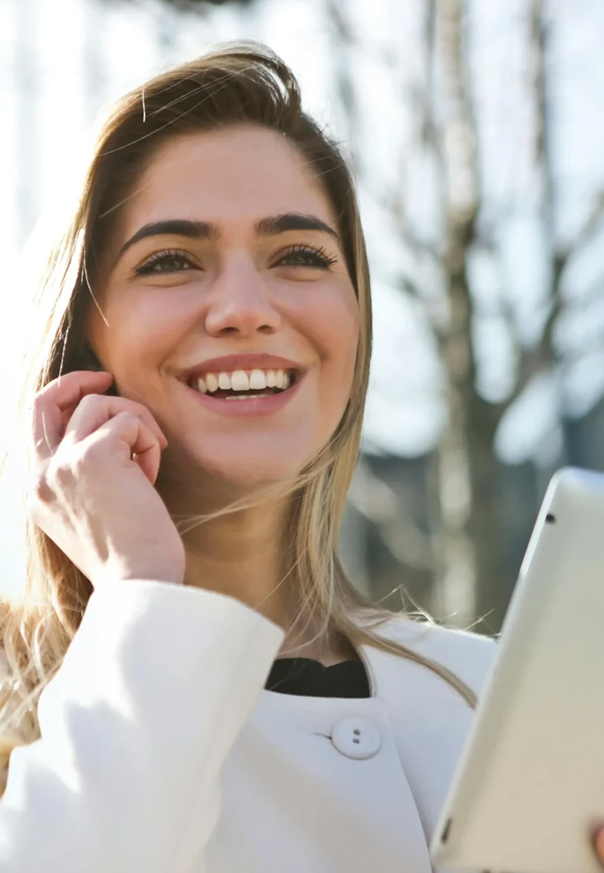 A woman with light brown hair and a white coat is smiling and holding a tablet. She appears to be outdoors.