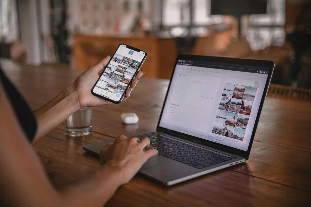 A person is shown working on a laptop, with a smartphone displaying a similar image grid next to it. A glass of water and wireless earbuds are also on the wooden table.