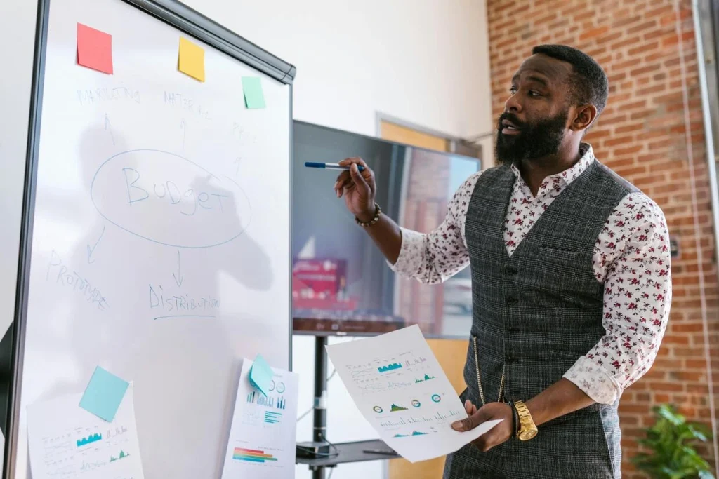 A man with a beard and a patterned vest is presenting at a whiteboard with the word "Budget" written on it. He's holding a marker and papers with charts.